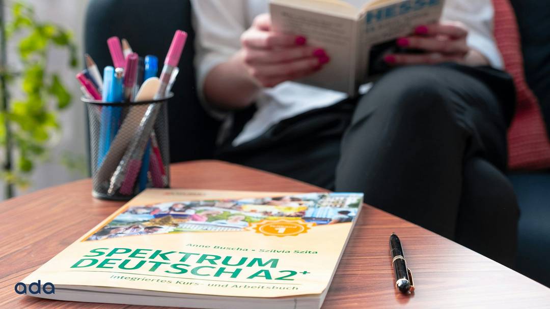 A German language learning book on a table with pens and with a person reading in the background.